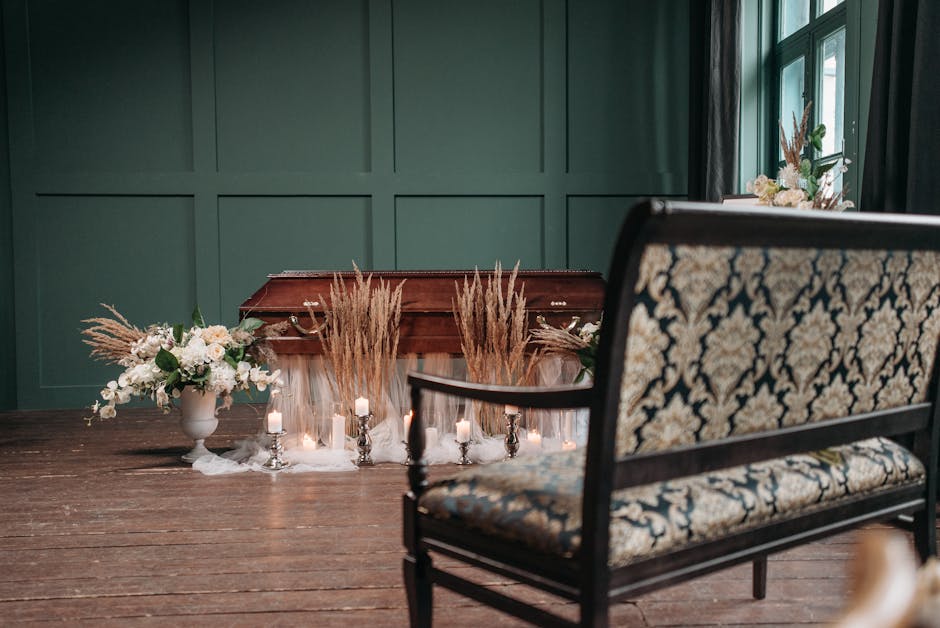 A wooden casket draped with white fabric is surrounded by floral arrangements and lit candles in silver holders, placed in a green-walled room with large windows. To the left, a white vase holds a bou