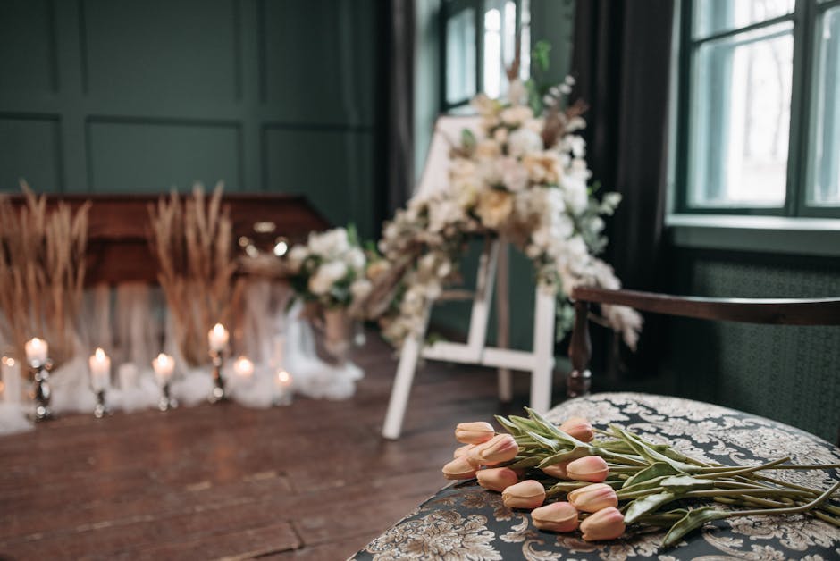 A floral arrangement setup featuring a bouquet of soft pink tulips with green stems and leaves resting on a patterned fabric surface in the foreground. In the background, there is a large, elaborate f