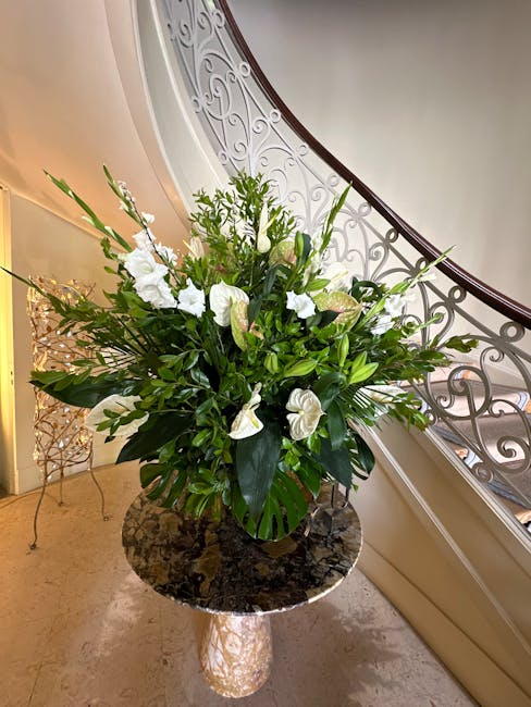 A large, lush floral arrangement featuring white lilies, anthuriums, and various green foliage, including broad and spiky leaves, all arranged in a round, dark green vase. The bouquet is placed on a p