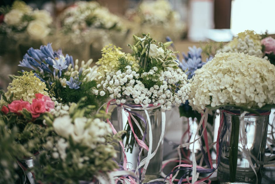 A variety of floral arrangements in glass vases featuring white hydrangeas, baby's breath, and pastel pink roses, accented with blue and yellow flowers, all set on a table with soft natural lighting. 